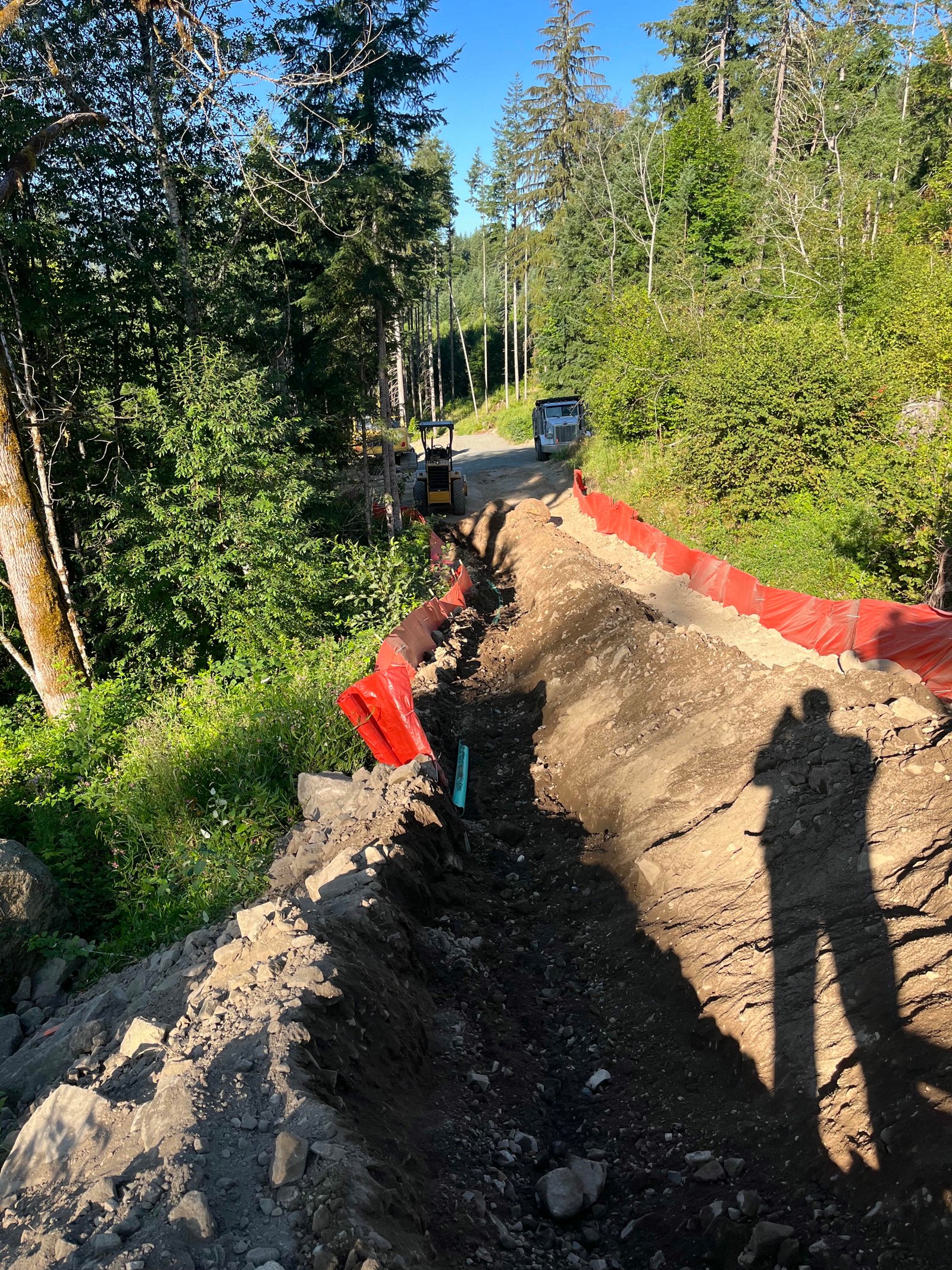 Deep utility trench excavation on a sloped property with safety fencing and heavy equipment in the background